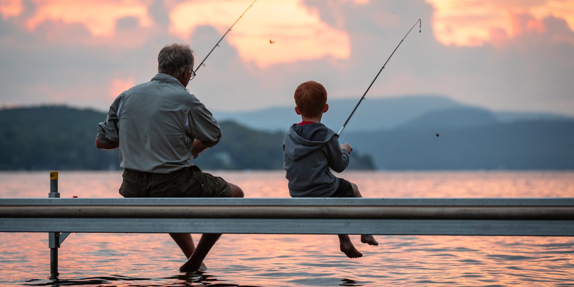 A grandfather is teaching his grandson to fish during summer. They are both sitting on the dock. It is a beautiful summer day at sunset. Across the lake, there is a mountain, Quebec, Canada.