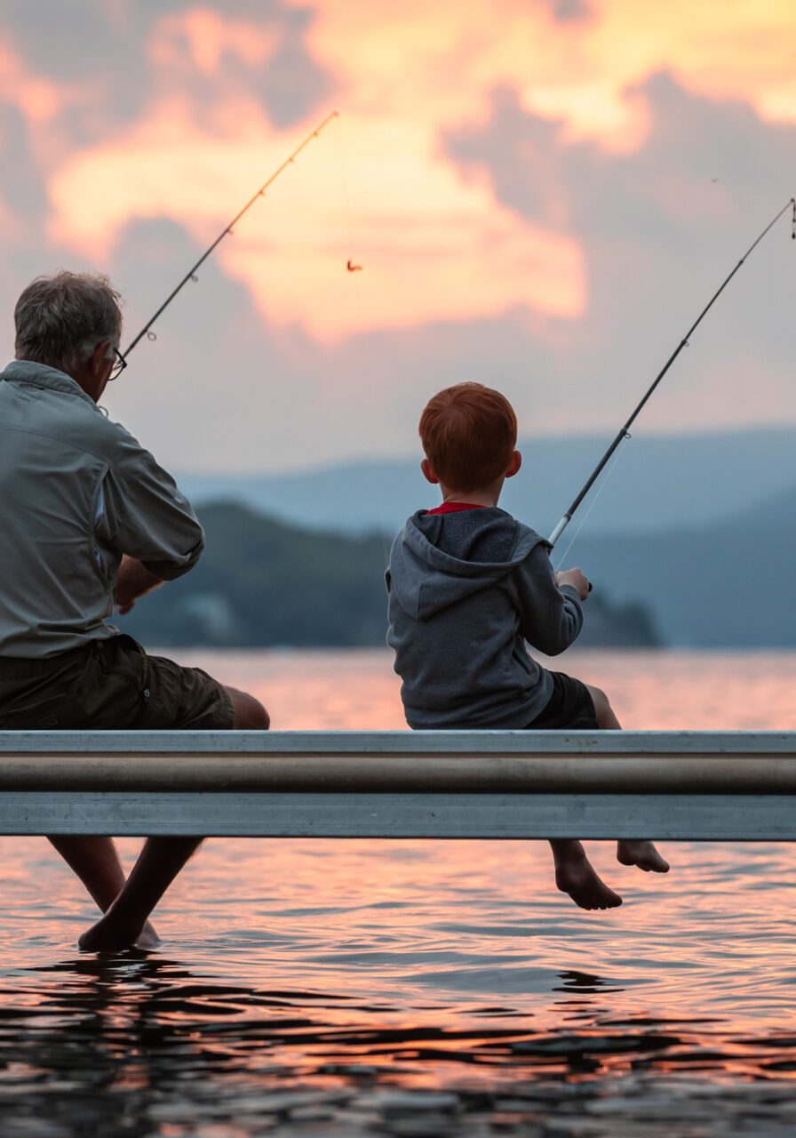 A grandfather is teaching his grandson to fish during summer. They are both sitting on the dock. It is a beautiful summer day at sunset. Across the lake, there is a mountain, Quebec, Canada.